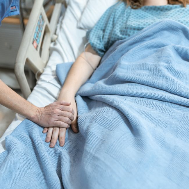 Female laying in a hospital bed with another person's hand on hers. This person is her enduring guardian that was appointed as part of her estate planning.