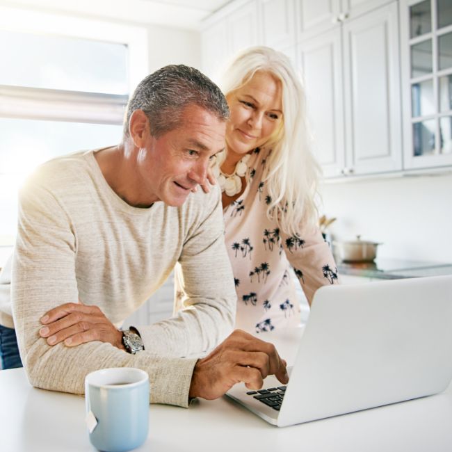 A mid-life couple standing in their white kitchen with a white laptop on the kitchen bench. They're reviewing their estate plan.